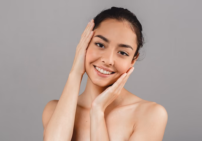 Smiling woman with dark hair holding her face with both hands against a gray background.
