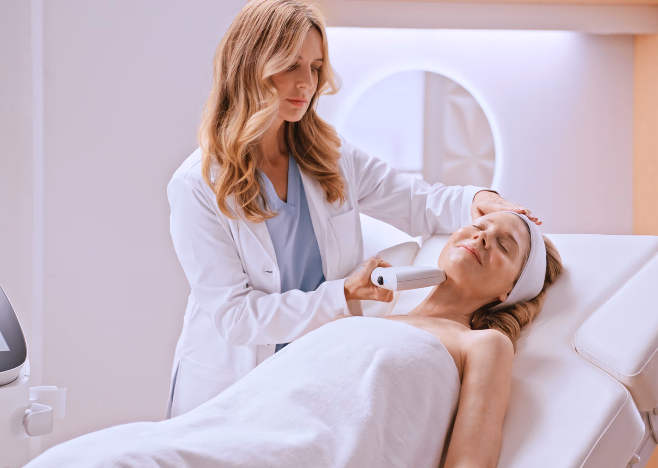 A female healthcare professional in a white coat performs a facial treatment with a handheld device on a relaxed woman lying on a treatment bed covered with a white towel.