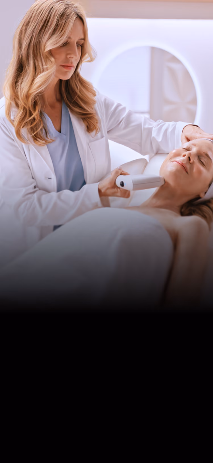 Blonde woman in a white coat performing a facial treatment on a lying woman using a handheld device in a bright, modern clinic.