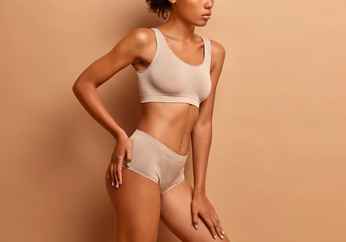 Woman with curly hair wearing beige seamless sports bra and shorts, posing sideways against a beige background.