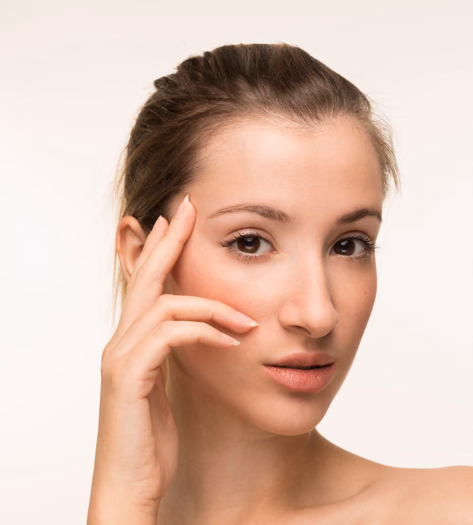 Close-up of a woman with clear skin touching her face gently with her fingers, looking directly at the camera against a plain background.