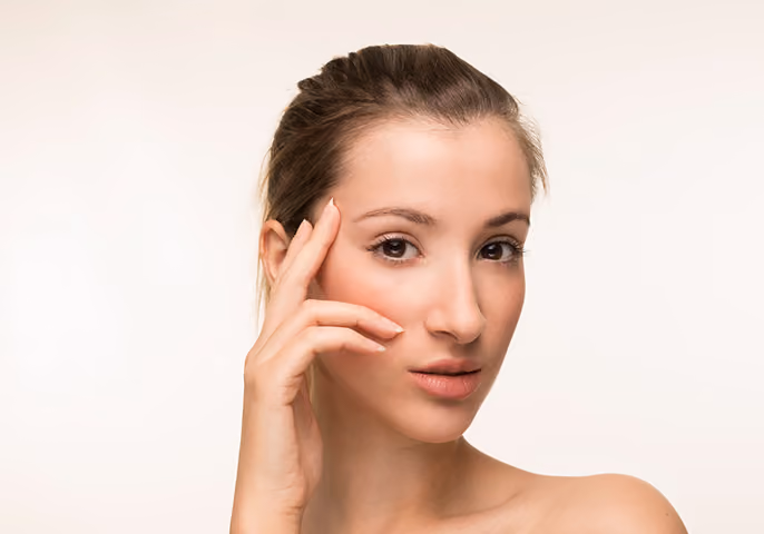 Portrait of a woman with light skin and brown hair pulled back, gently touching her face with her hand against a plain light background.