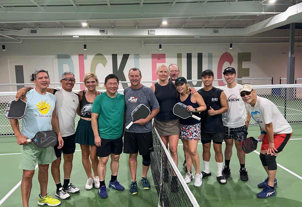 Group of ten people standing together on an indoor pickleball court holding paddles behind the net.