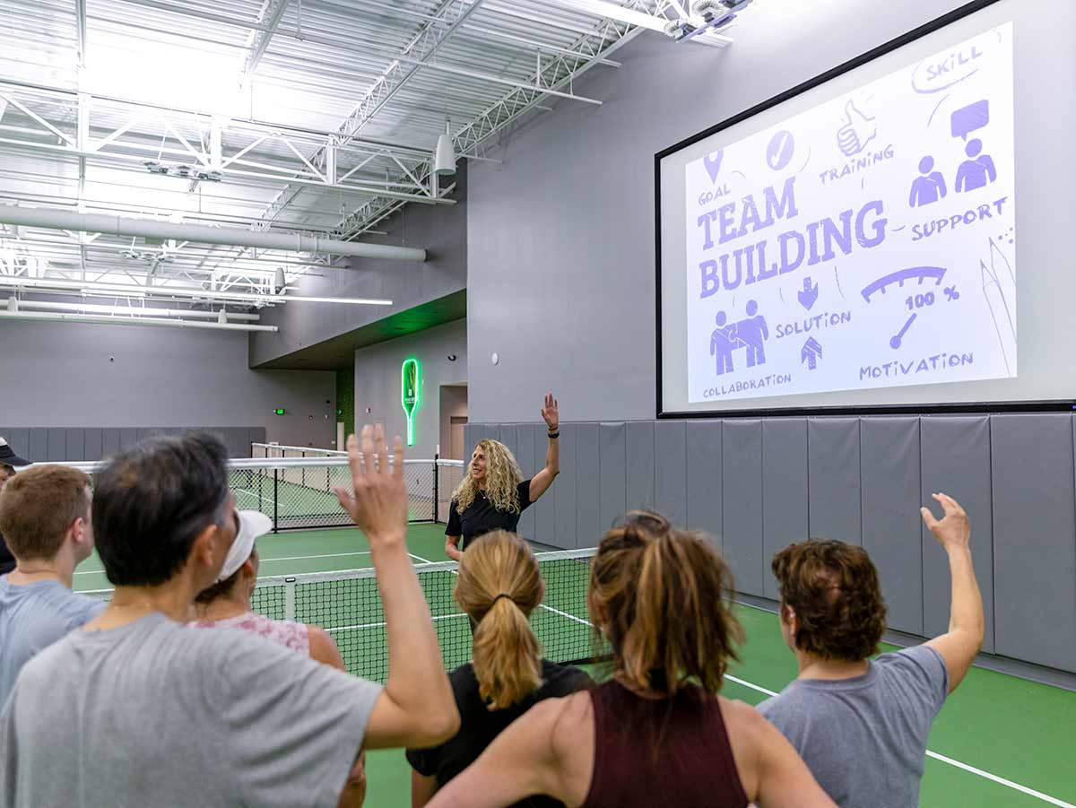 Instructor leading team building session with group raising hands in indoor sports court.