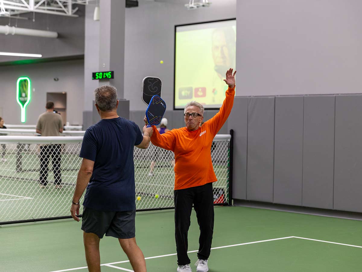 Two men playing pickleball indoors, one in orange shirt raising arm, the other in navy shirt facing away.