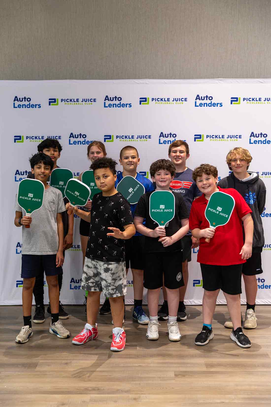 Group of nine kids holding green Pickle Juice pickleball paddles in front of sponsor backdrop.