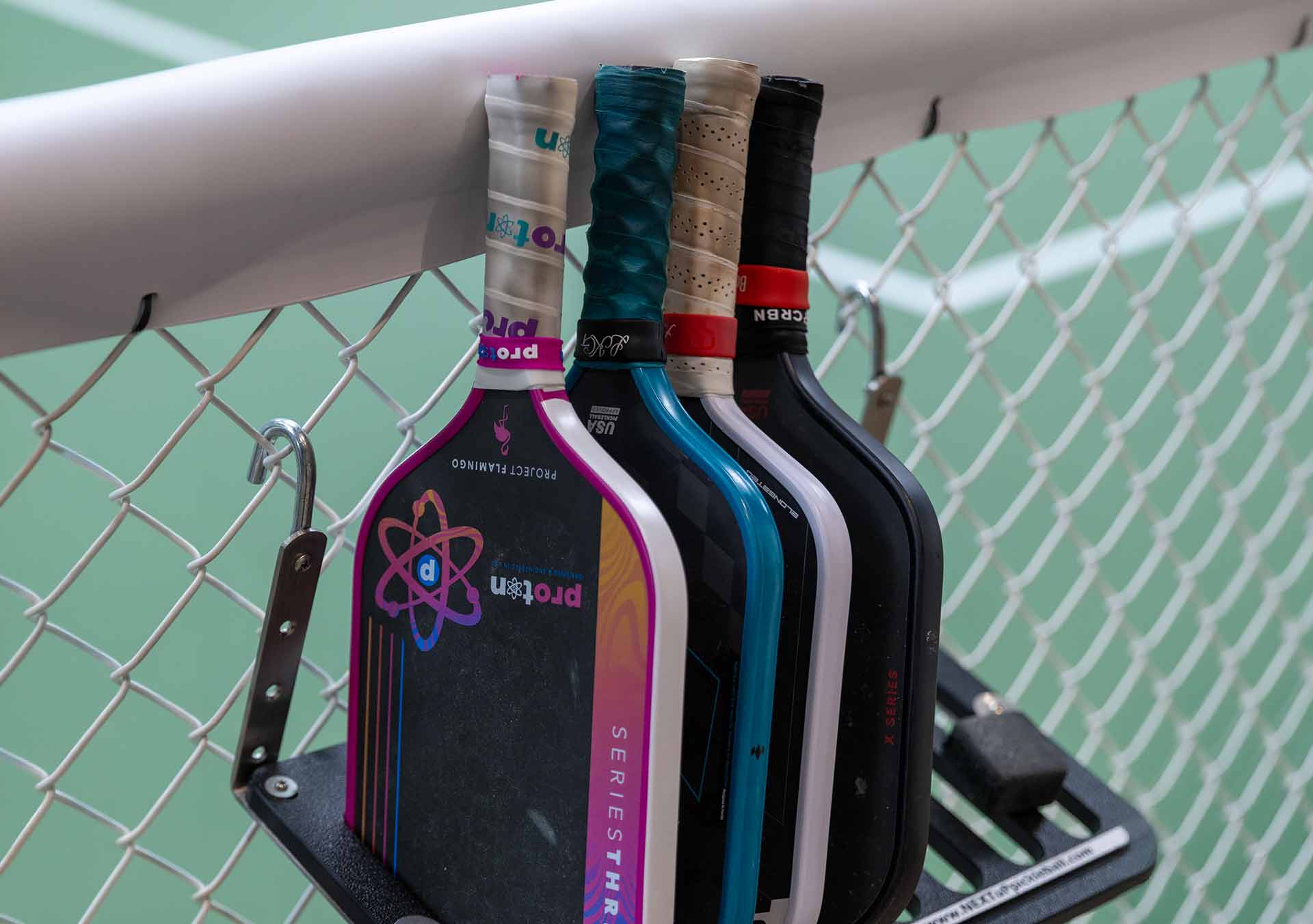 Four pickleball paddles with wrapped handles hanging on a metal rack against a chain-link fence.
