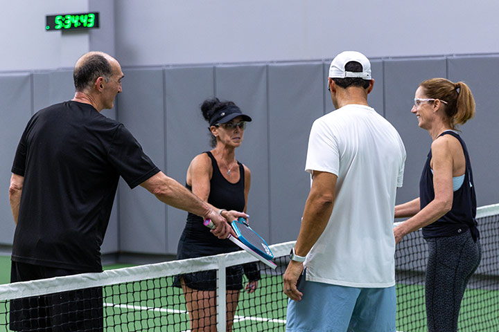 Four adults standing on a pickleball court holding paddles and talking near the net indoors.