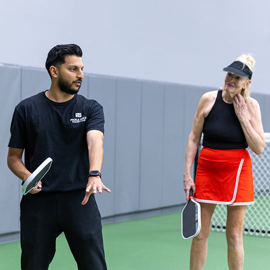 Man coaching an elderly woman in pickleball on an indoor court, both holding paddles.