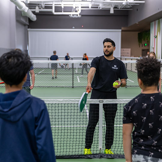 Man holding a paddle and ball teaching pickleball to young players on an indoor court.