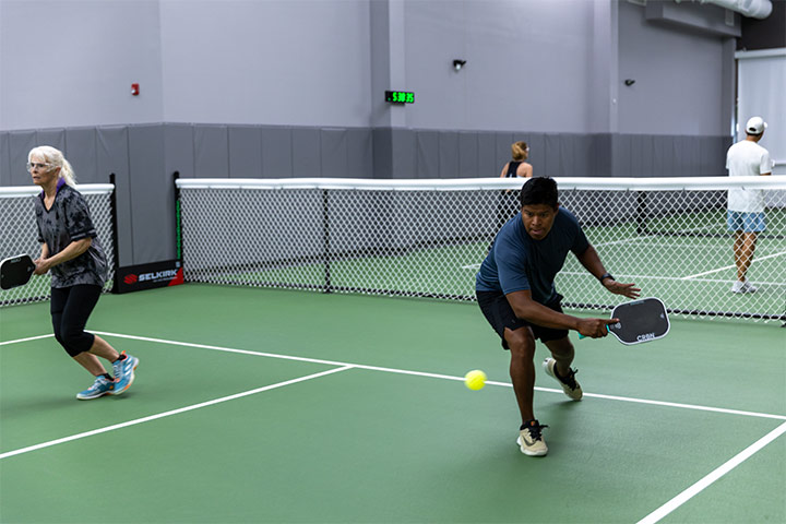 Two people playing pickleball indoors, one about to hit the yellow ball with a paddle.