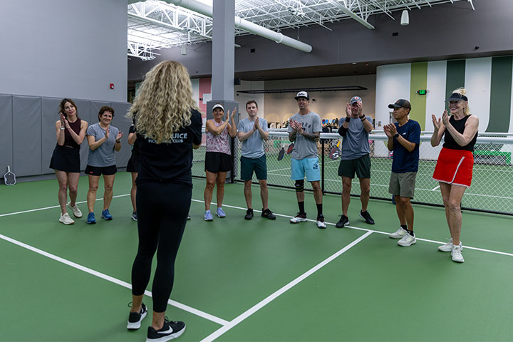 Group of pickleball players clapping while a woman in black faces them on an indoor court.