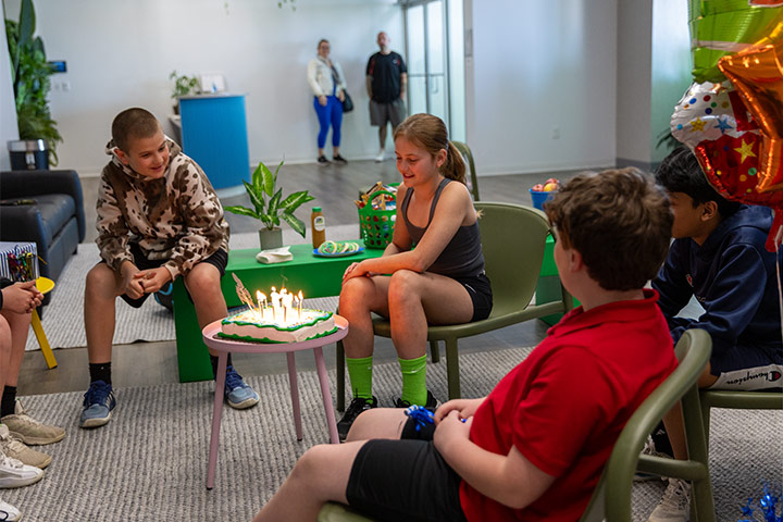 Children gathered around a birthday cake with lit candles in a casual indoor setting.
