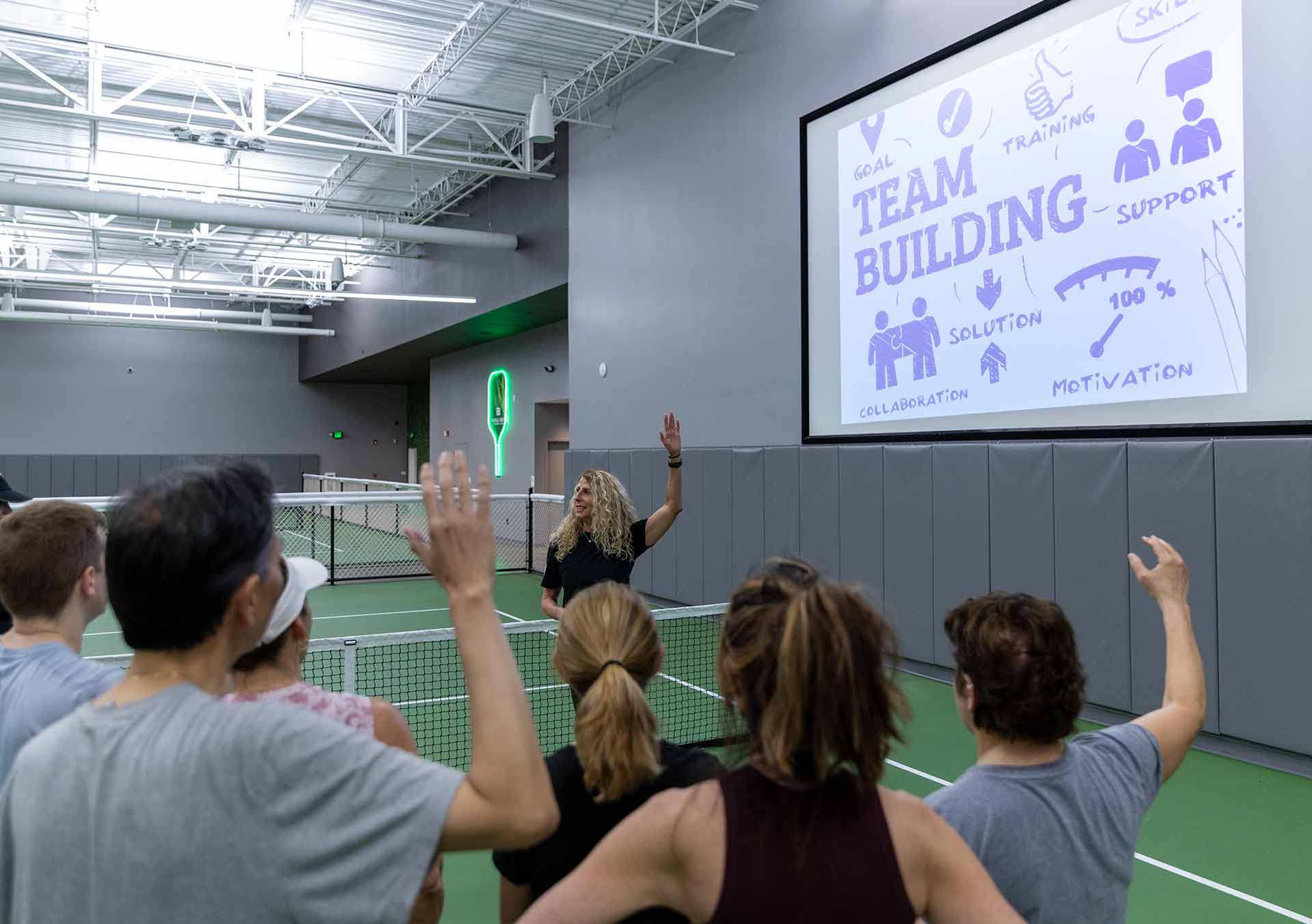 Instructor leads a team-building session on an indoor pickleball court with participants raising hands.