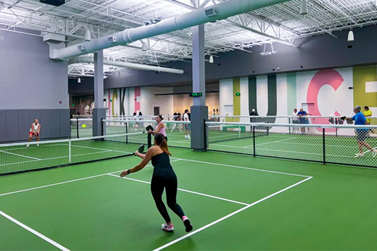 Several people playing pickleball indoors on green courts separated by fencing and large colorful letters on walls.