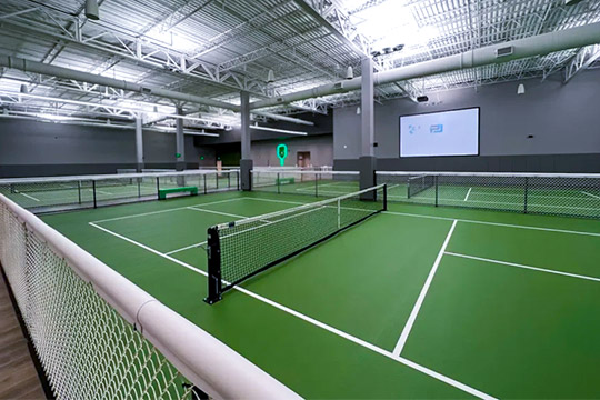 Indoor pickleball courts with green floors, white lines, and black nets under a metal roof.