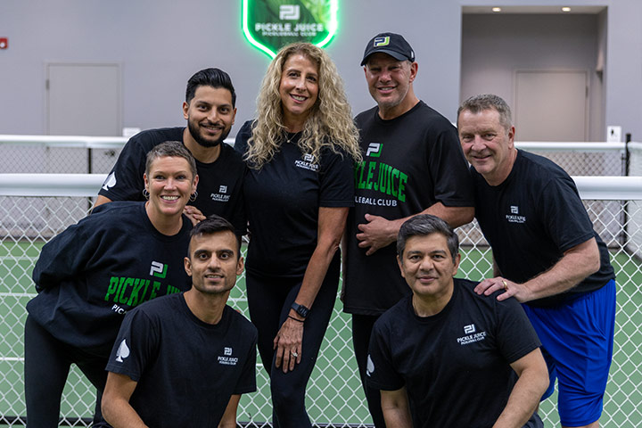 Seven smiling adults posing on an indoor pickleball court wearing Pickle Juice club shirts.
