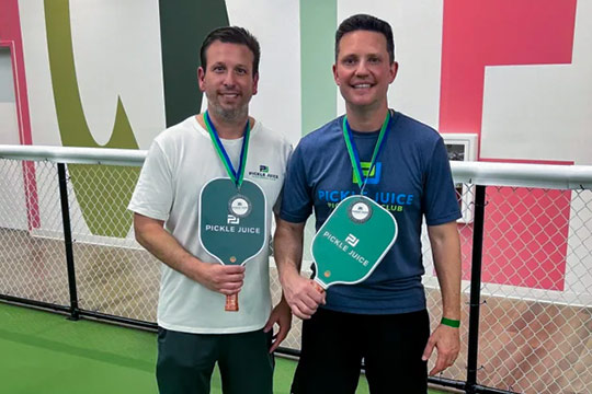 Two men holding Pickle Juice pickleball paddles, wearing medals, standing on a pickleball court.