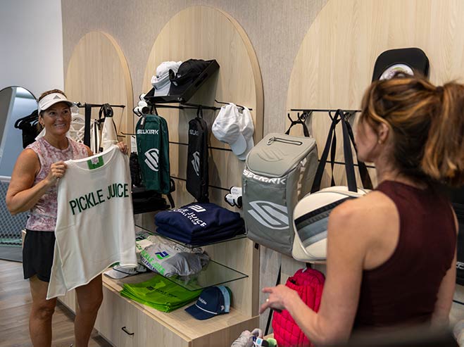 Two women shop for pickleball apparel and gear, one holding a white 'PICKLE JUICE' shirt in a store.