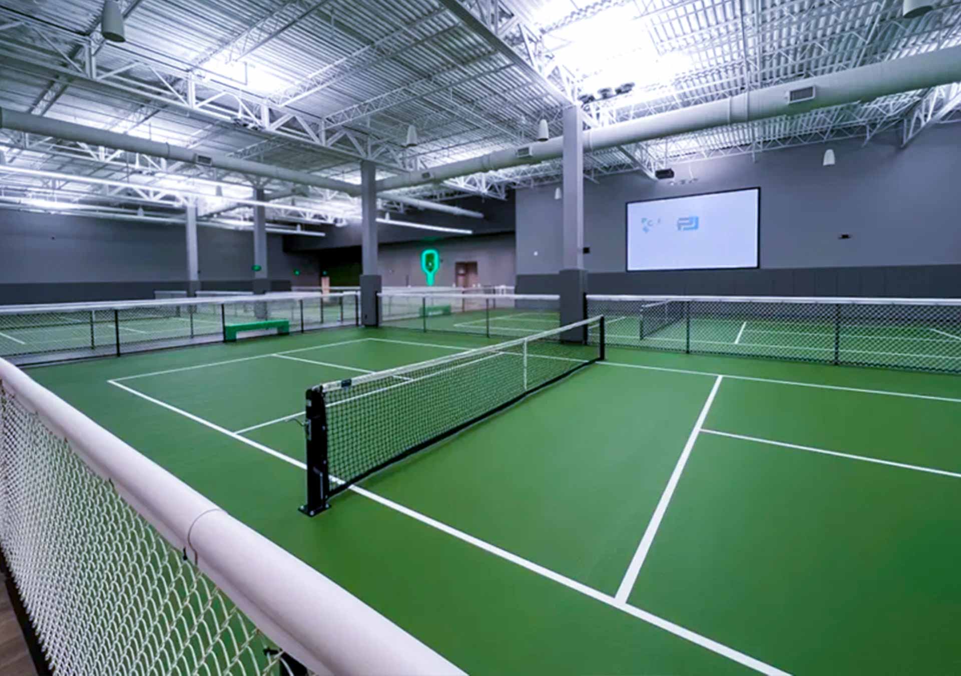 Indoor green pickleball courts with white boundary lines and nets under bright industrial lighting.