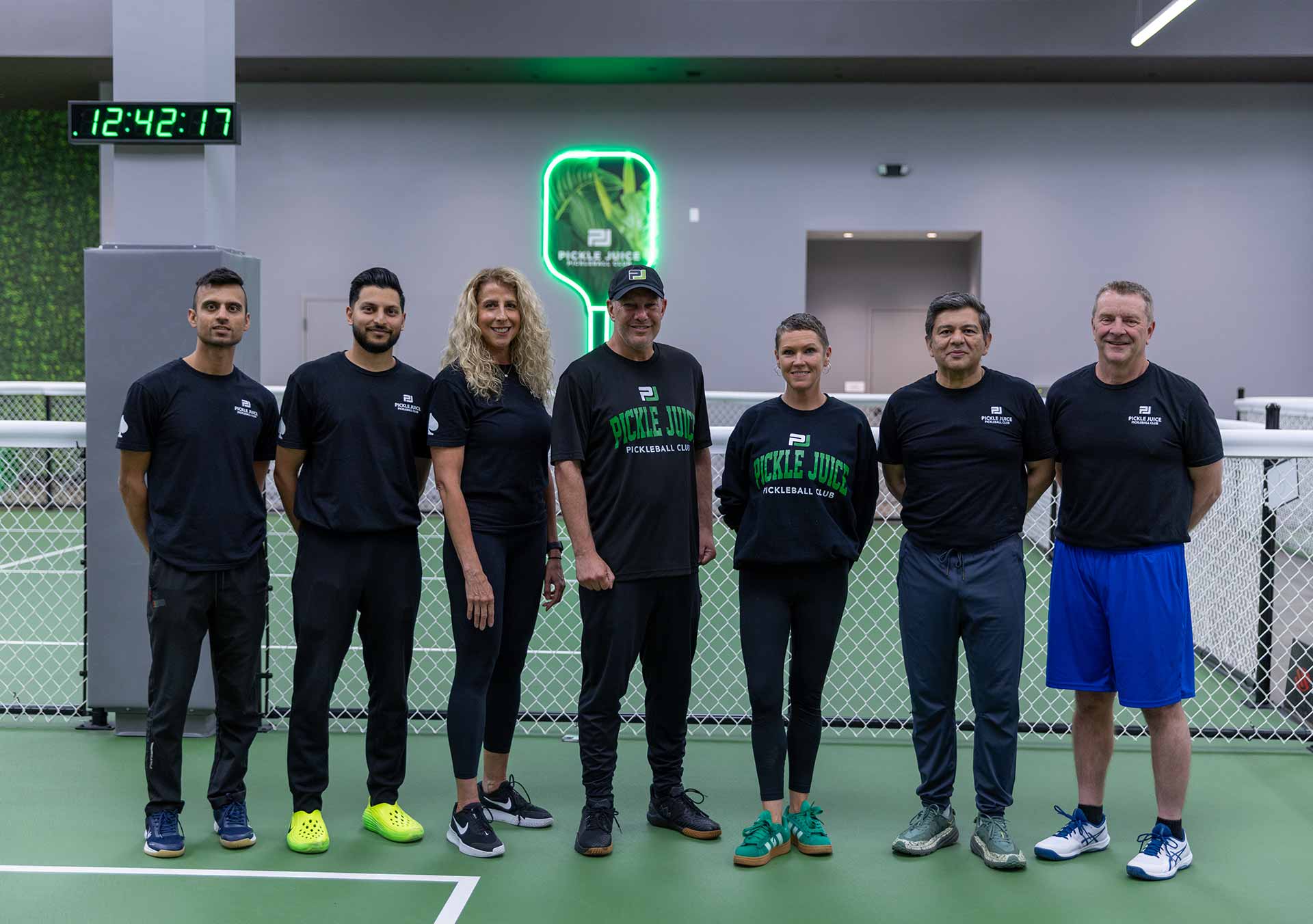 Seven people standing on an indoor pickleball court wearing Pickle Juice Pickleball Club apparel.