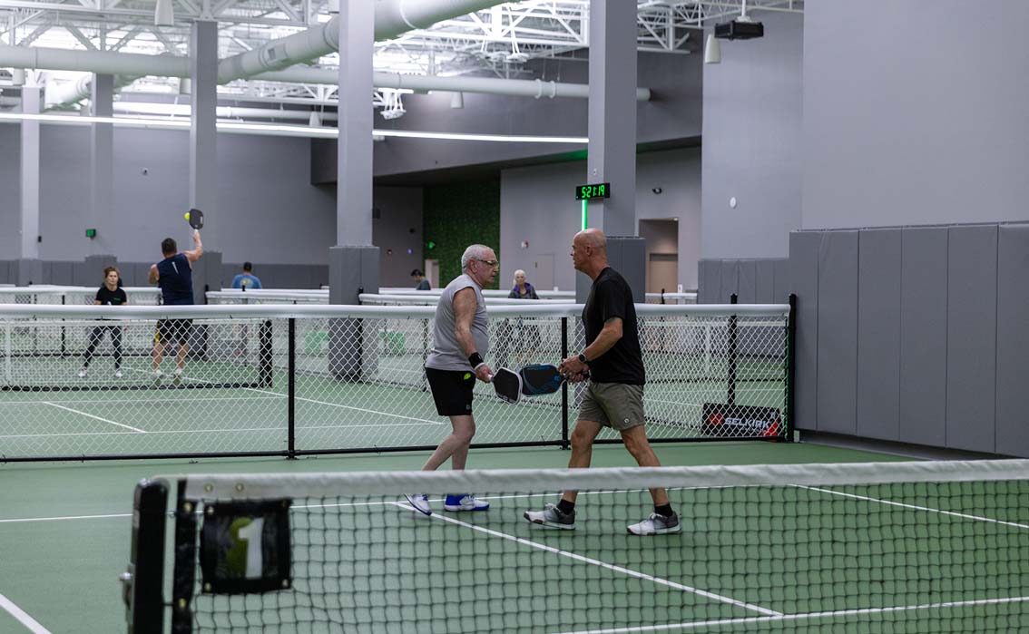 Two older men playing pickleball on an indoor court, holding paddles and facing each other.