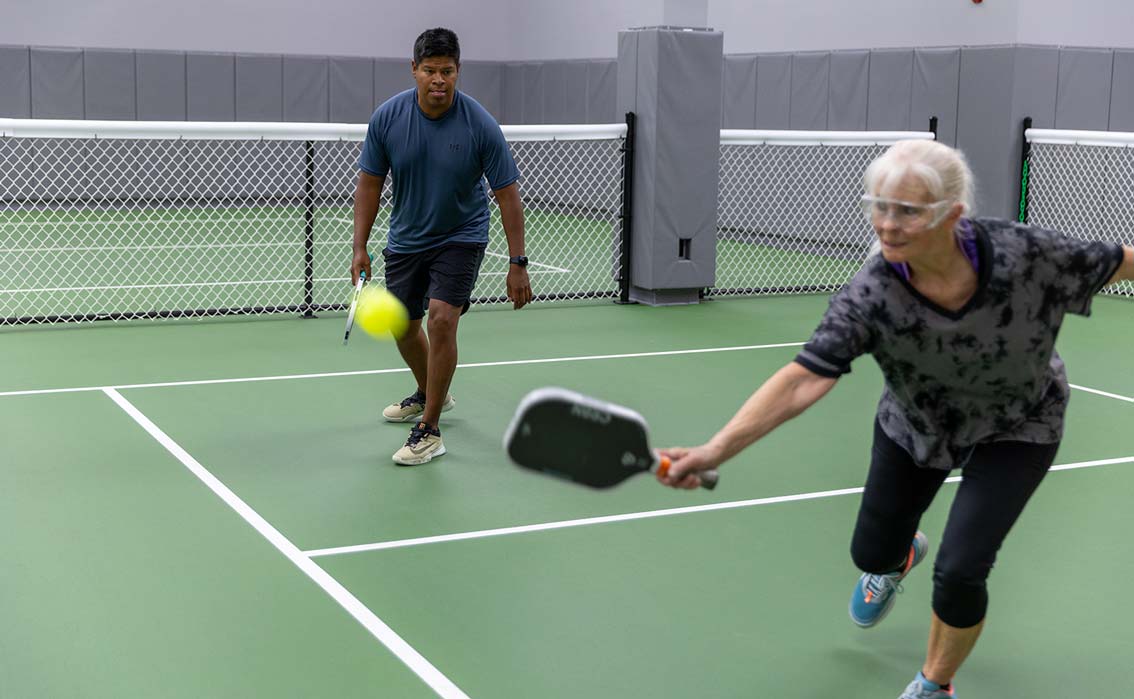 Older woman lunges to hit a yellow pickleball with a paddle as a man watches on an indoor court.