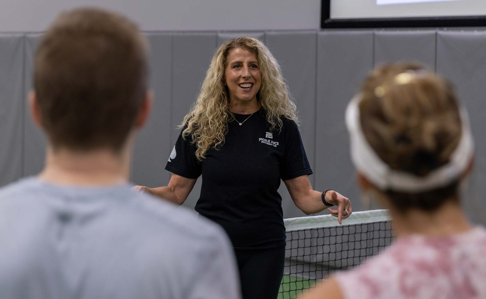 Woman with curly hair in a black Pickle Juice shirt speaking to two people near a pickleball net indoors.