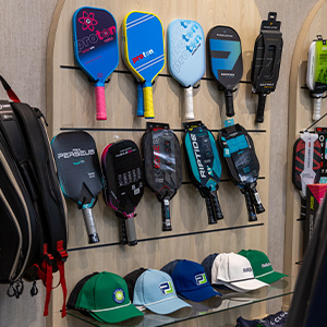 Store display of colorful pickleball paddles and caps on a wood wall and glass shelves.