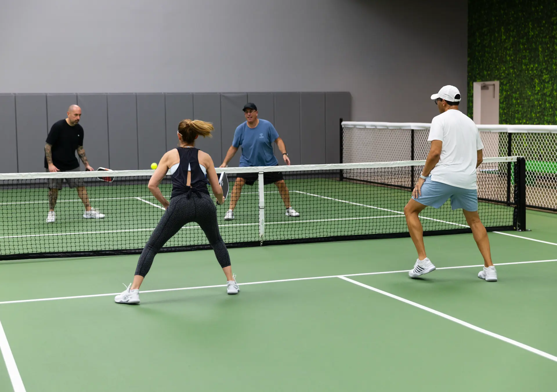 Four adults playing doubles pickleball indoors on a green court with a yellow ball in play.