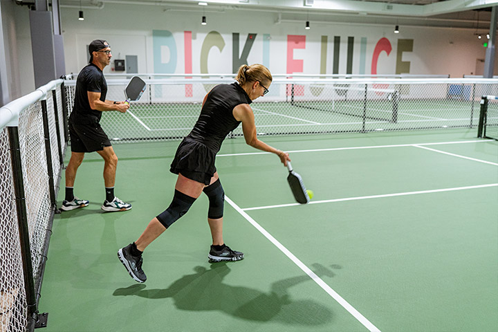Two players in black outfits playing pickleball on an indoor court with PICKLEJUICE on the wall.