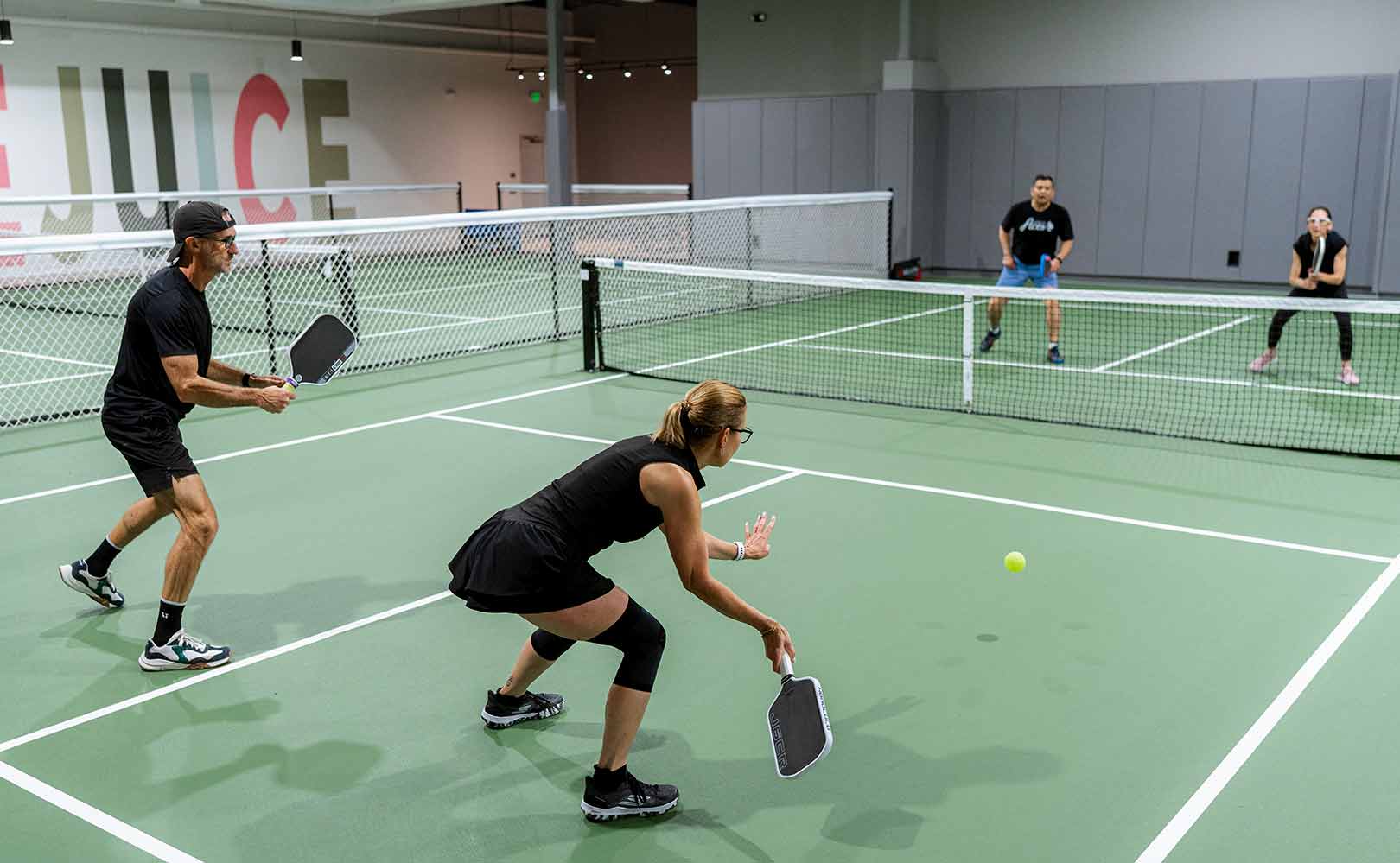 Four adults playing doubles pickleball on an indoor court, two players ready to hit the ball.