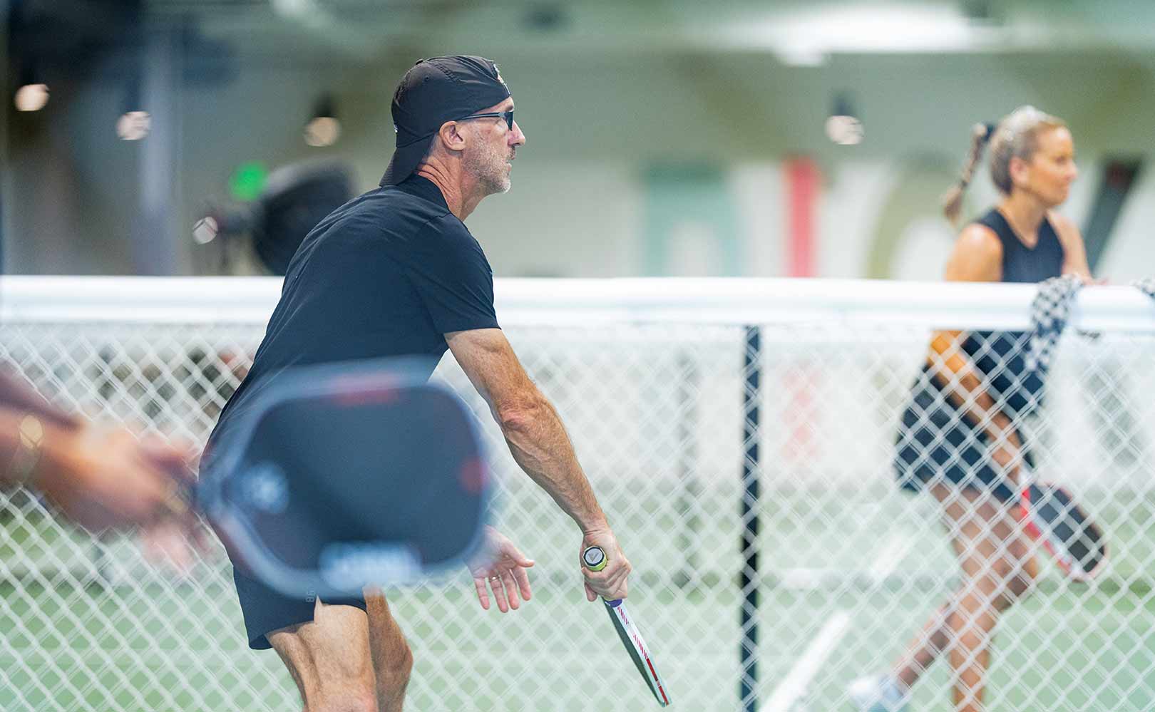 Man and woman playing pickleball indoors with paddles and ball near the net.