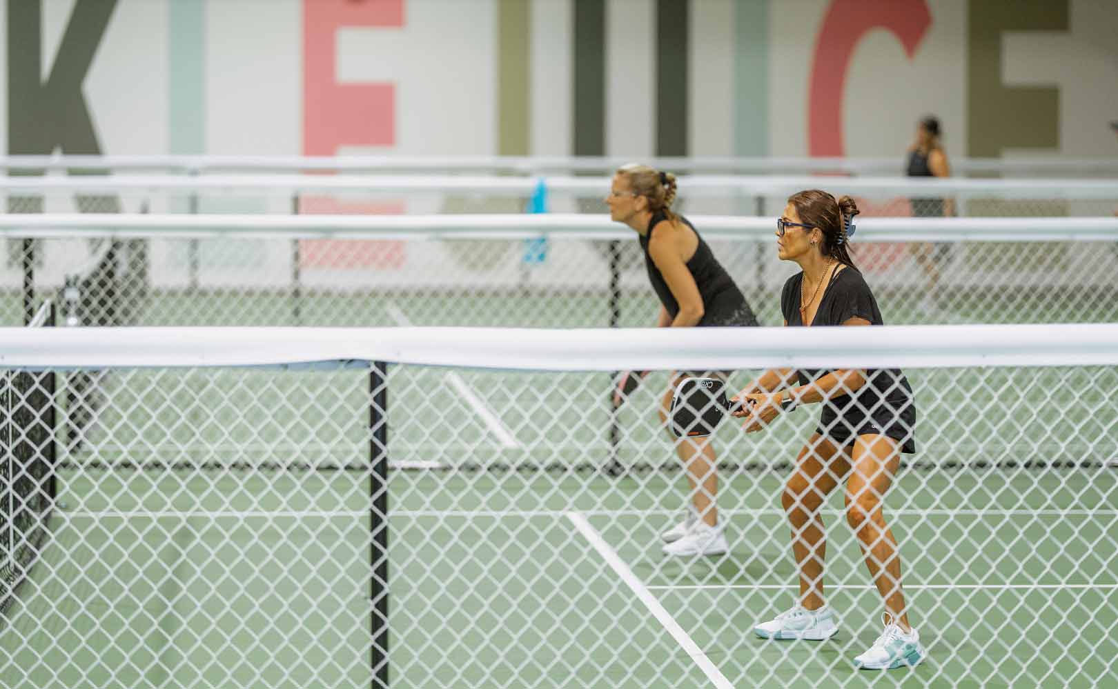 Two women in athletic wear playing pickleball indoors, holding paddles and ready to hit the ball.