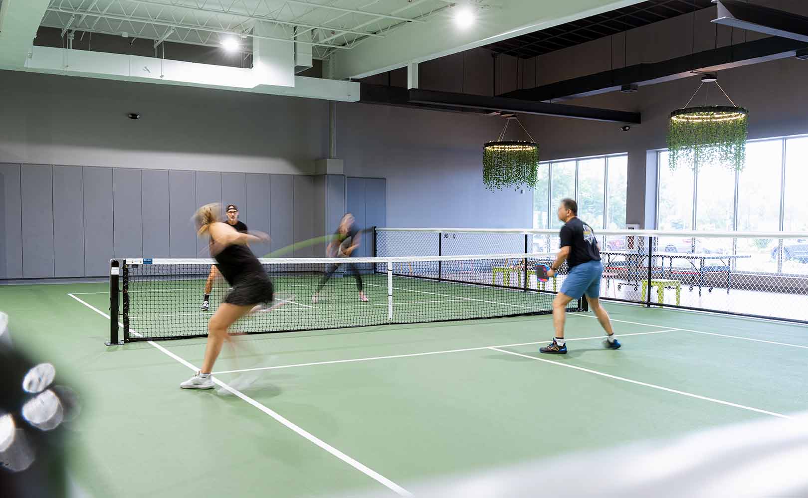 Four people playing doubles pickleball indoors on a green court with hanging light fixtures.