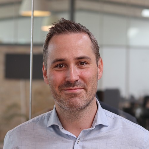 Smiling man with short brown hair and light beard wearing a light blue checkered shirt in an office setting.