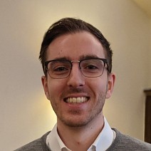 Smiling man with glasses, short dark hair, and wearing a grey sweater over a white collared shirt against a plain light background.
