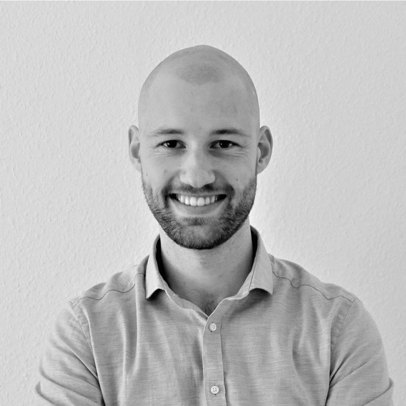Smiling bald man with a beard wearing a buttoned-up shirt, standing against a plain wall.