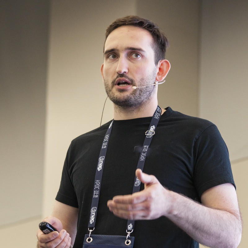 Man with short dark hair and beard wearing a black t-shirt and a conference lanyard is speaking with a headset microphone and gesturing with his hands.