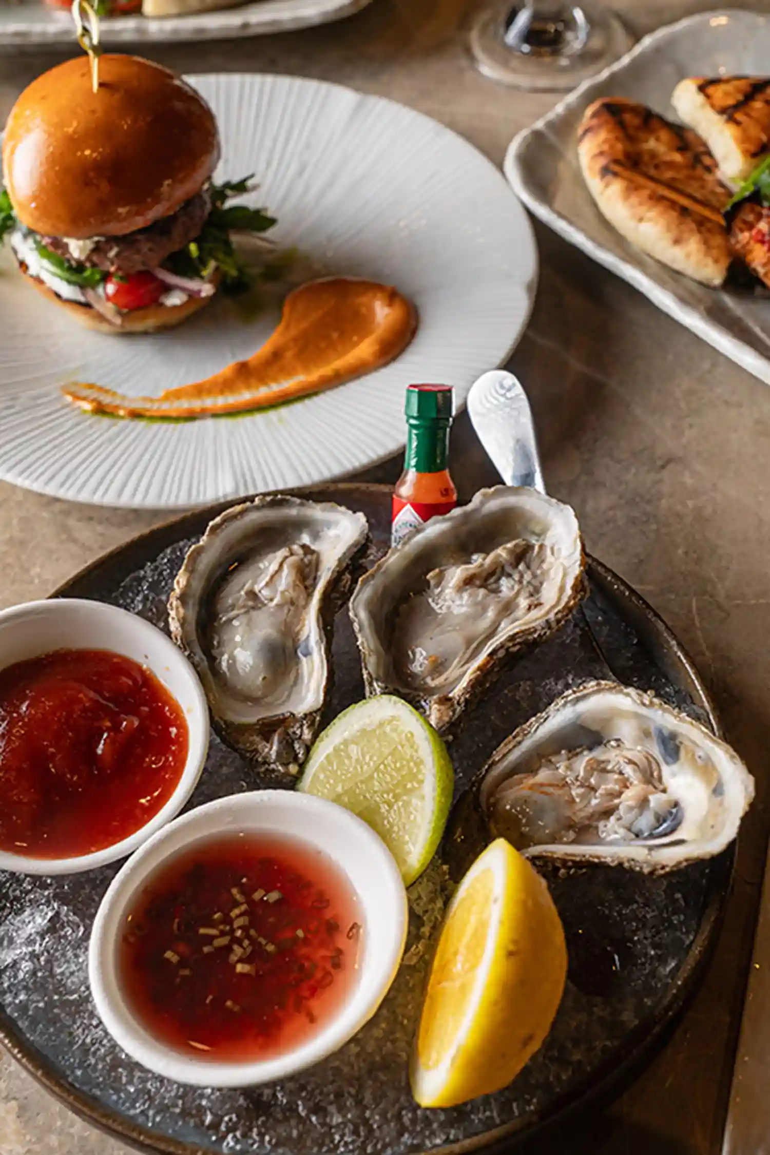 A platter of fresh oysters served on ice with dipping sauces and citrus at Ziva Miami, accompanied by a burger in the background