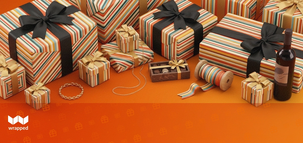 Gold bracelet and chain necklace beside a small chocolate box, ribbon spool and wine bottle on a warm amber surface