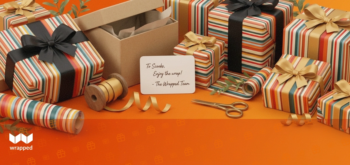 Kraft gift box, patterned wrapping paper roll, satin ribbon, brass scissors and a handwritten card on a warm amber table