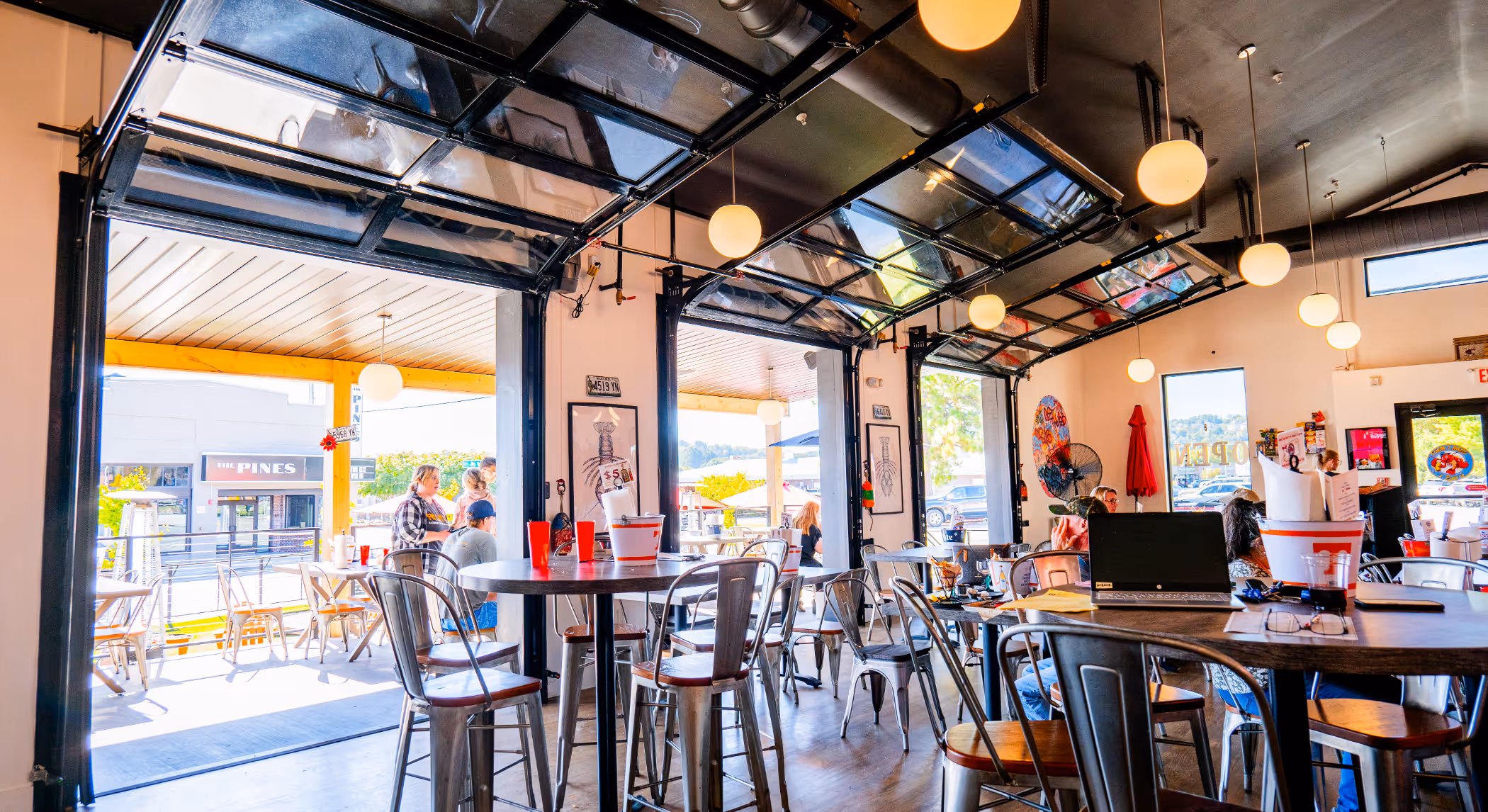 Bright restaurant interior with large open garage doors, metal chairs, round tables, hanging globe lights, and patrons dining inside and on the patio.