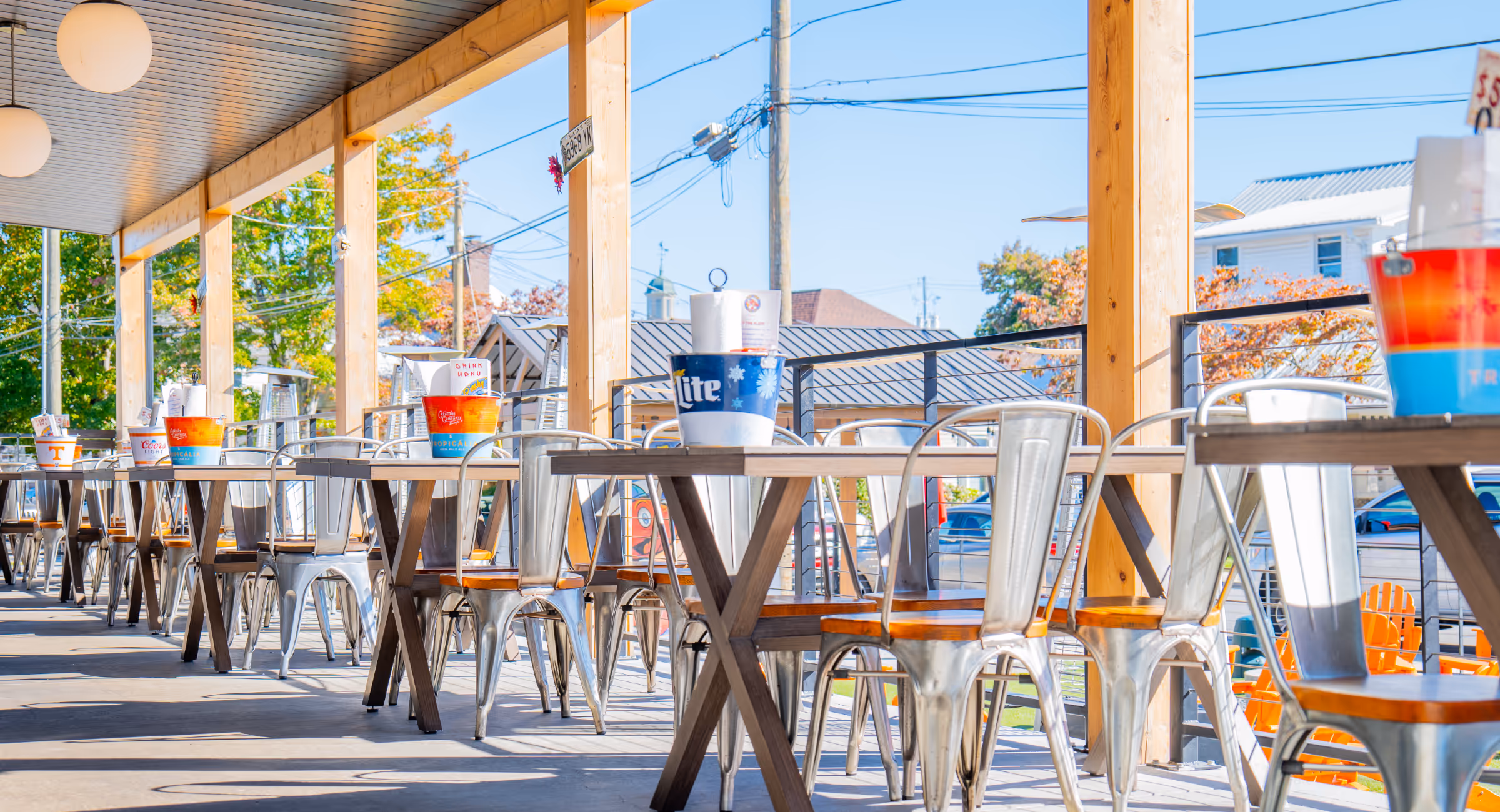 Outdoor covered patio with metal and wood chairs and tables, each table holding branded drink buckets and napkins.