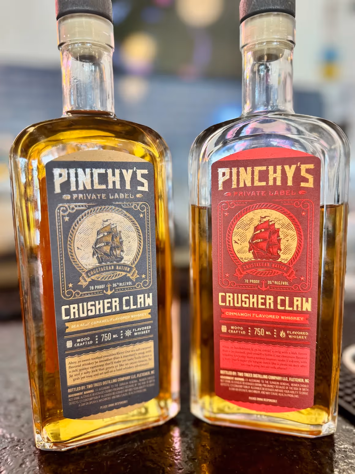 Two bottles of Pinchy's Crusher Claw whiskey, one sea salt caramel flavored with a black label and one cinnamon flavored with a red label.