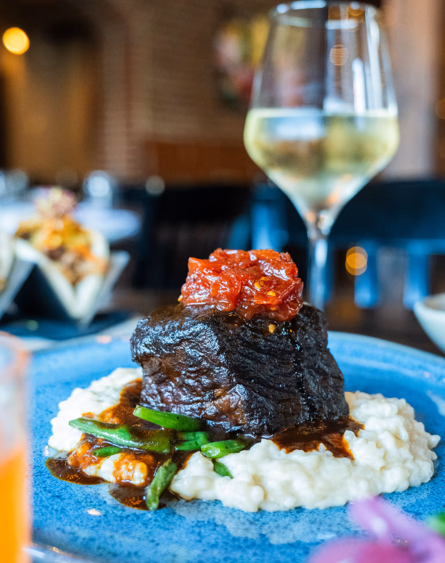 Plate with braised beef topped with tomato relish, served on mashed potatoes and green beans, with a glass of white wine in the background.