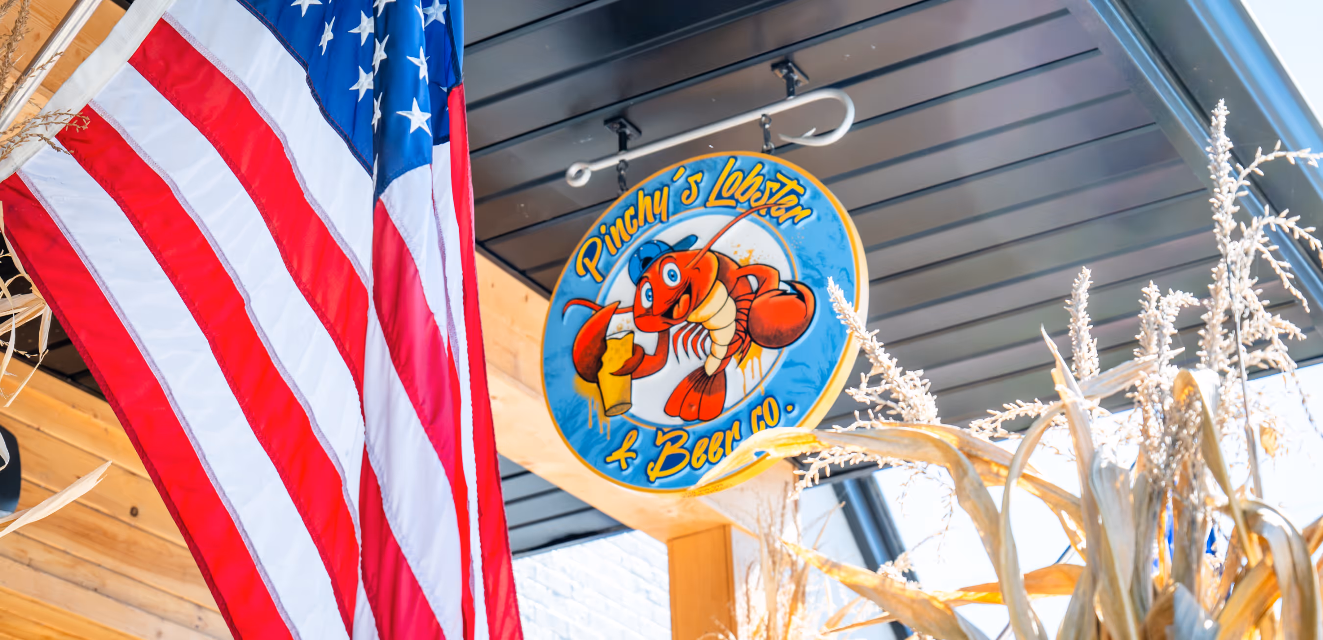 Sign for Pinchy's Lobster & Beer Co. featuring a cartoon lobster holding a beer glass, alongside a hanging American flag.