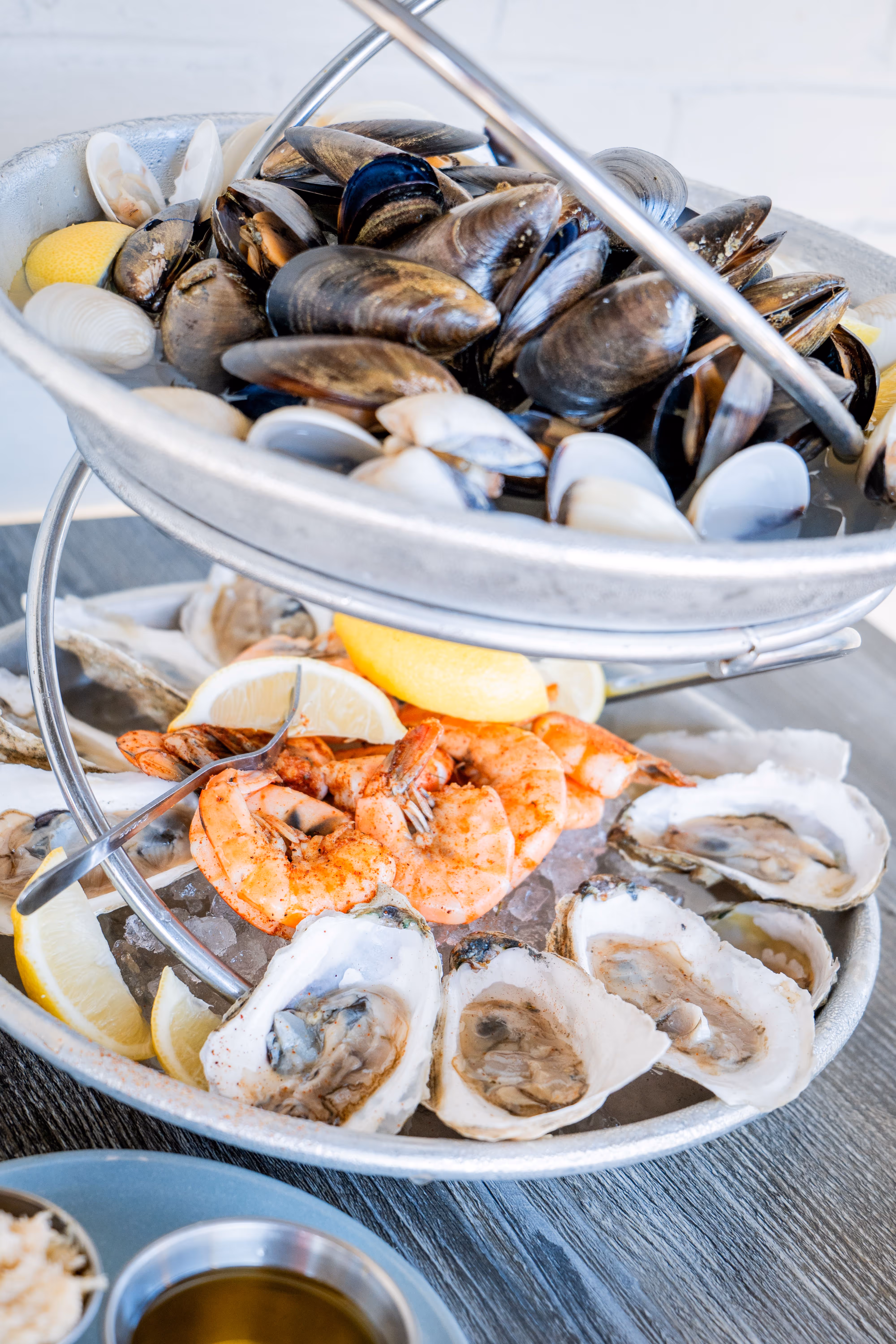 Two-tier seafood platter with cooked shrimp, oysters on ice, mussels, clams, and lemon wedges.