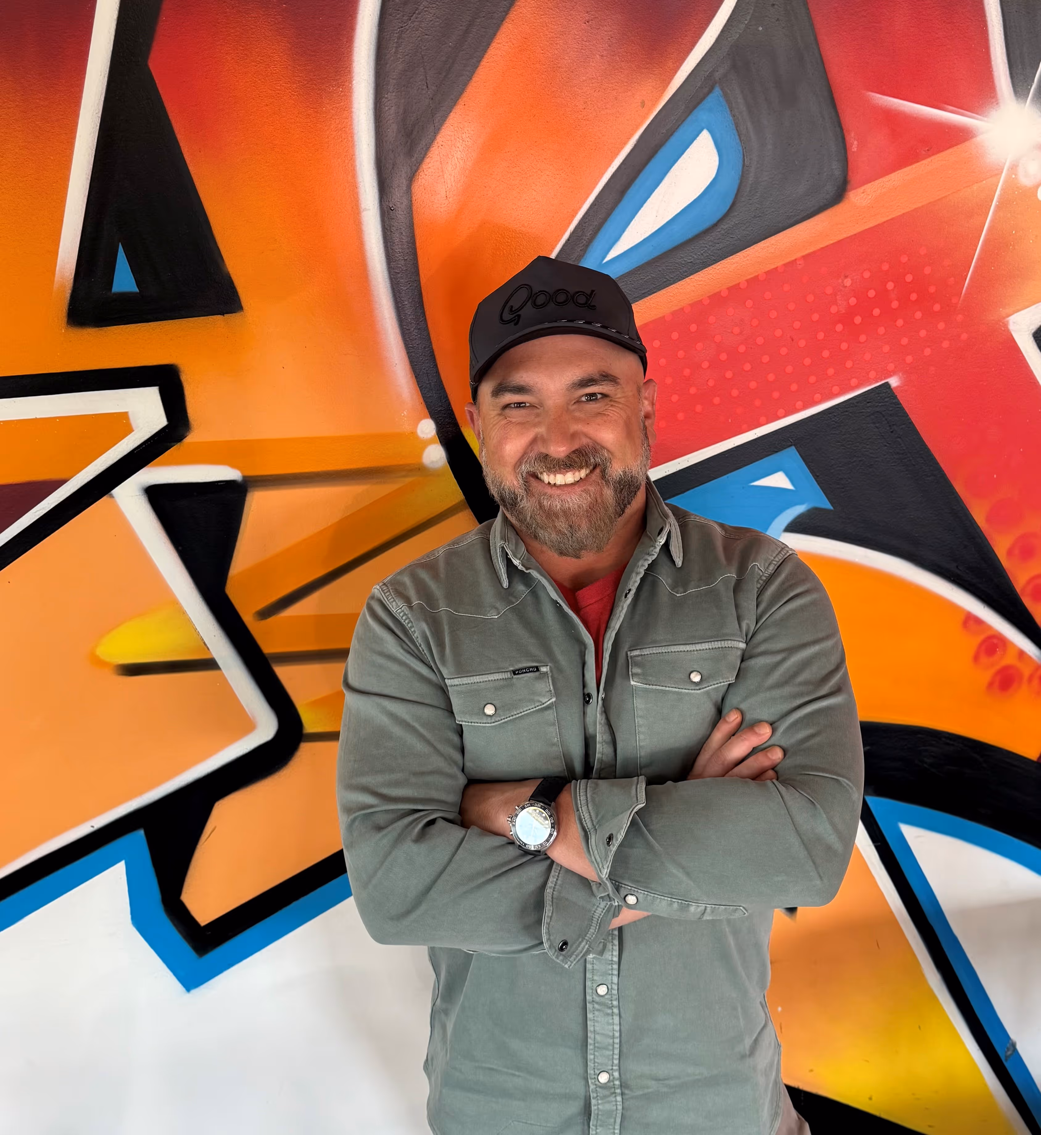 Smiling man with beard, green shirt, black cap standing with arms crossed in front of colorful graffiti wall.