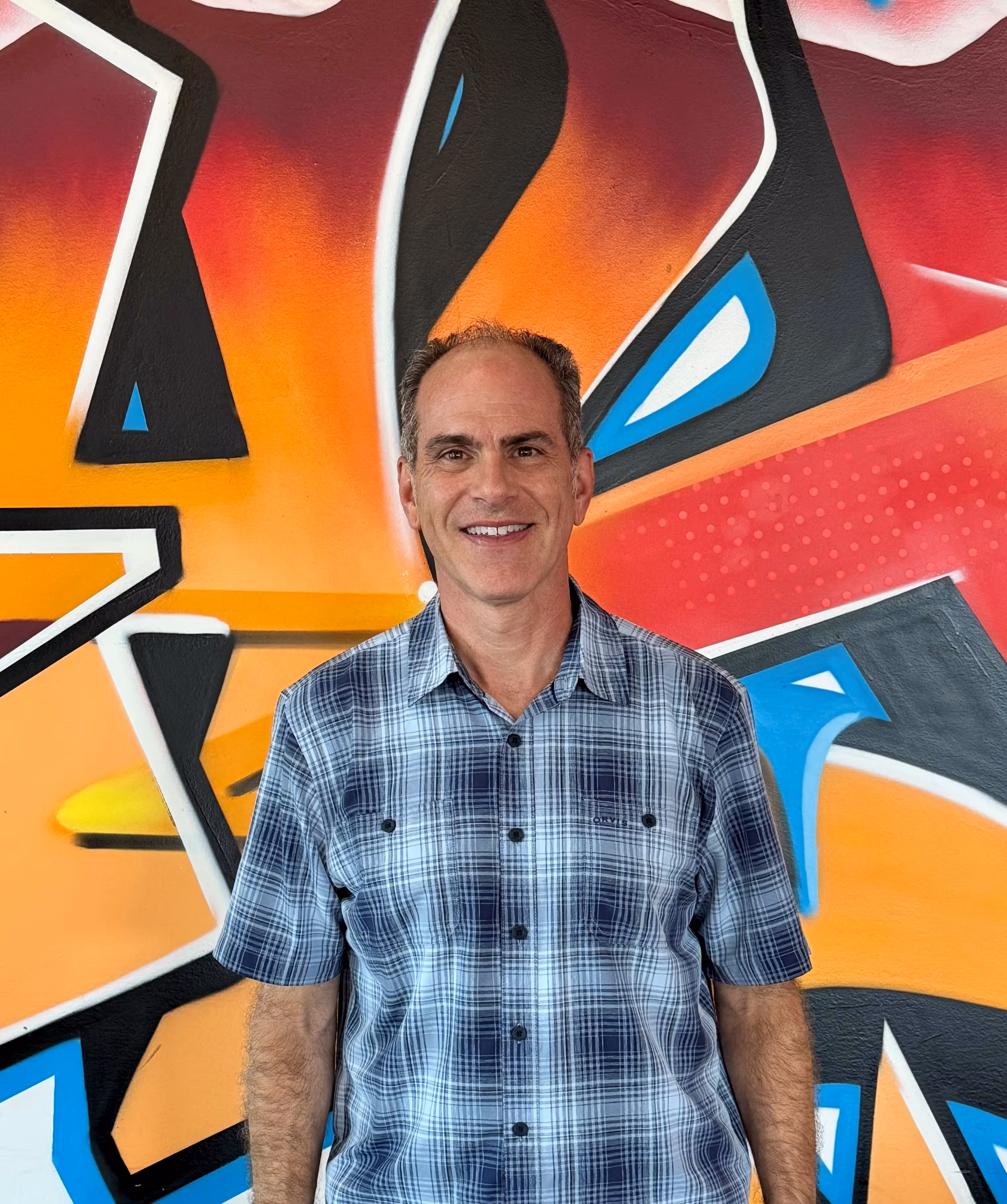 Smiling man in blue plaid shirt standing in front of colorful abstract graffiti wall.